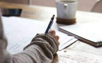 Eine Person in einem Wollpullover sitzt an einem brauen Holztisch und füllt mit einem Kugelschreiber ein Formular aus.